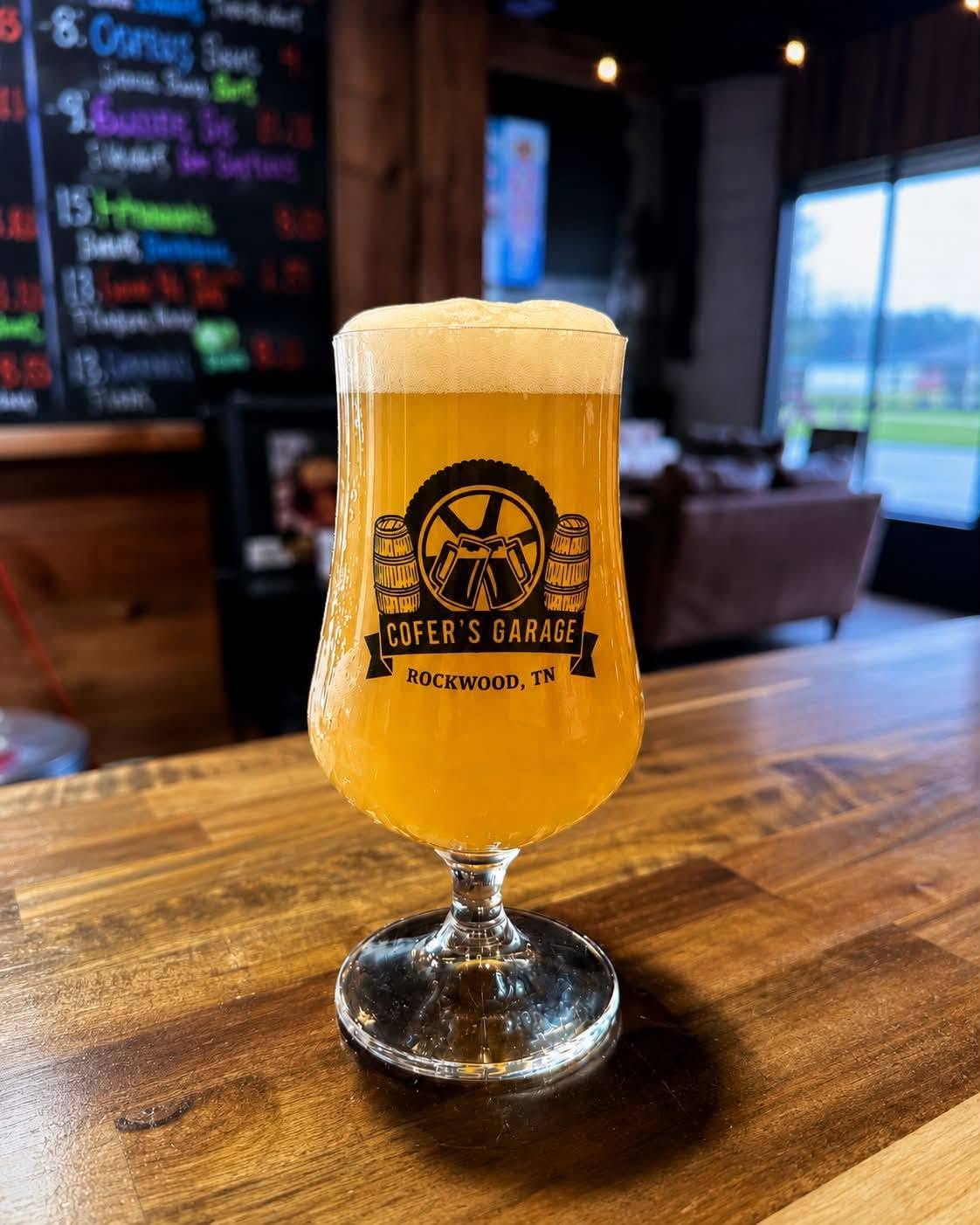 A branded Cofer's Garage beer glass filled with golden ale on the wooden bar